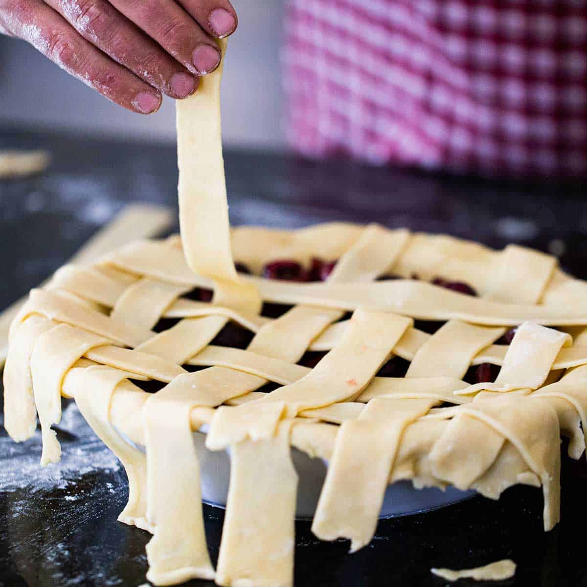A person braiding strips of pie dough over the top of an uncooked cherry pie to from a lattice formation.