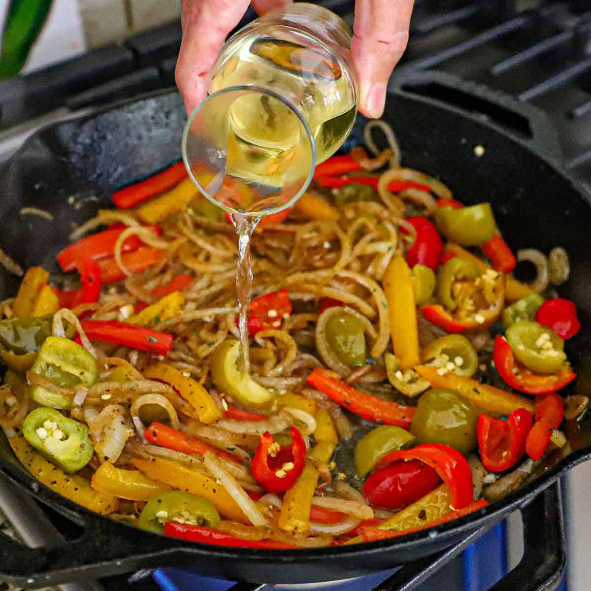 A person pouring white wine from a small carafe into a large cast-iron skillet filled with simmering sautéed onions, strips of bell peppers, and chopped pickled cherry peppers.