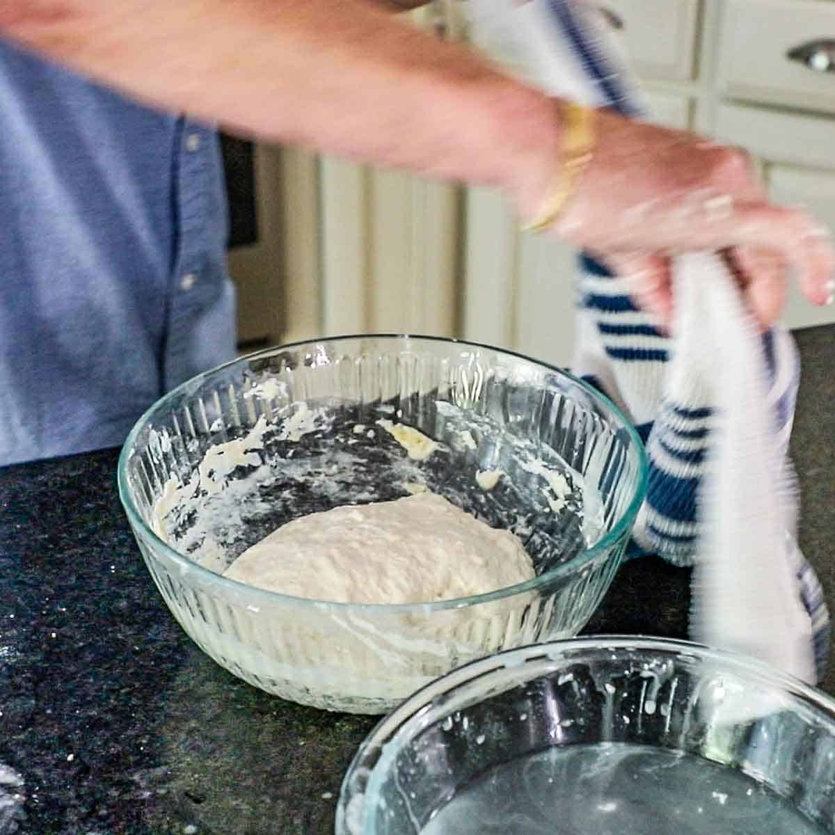 A person placing a kitchen towel over a large glass bowl that contains a mound of partially risen ciabatta dough.