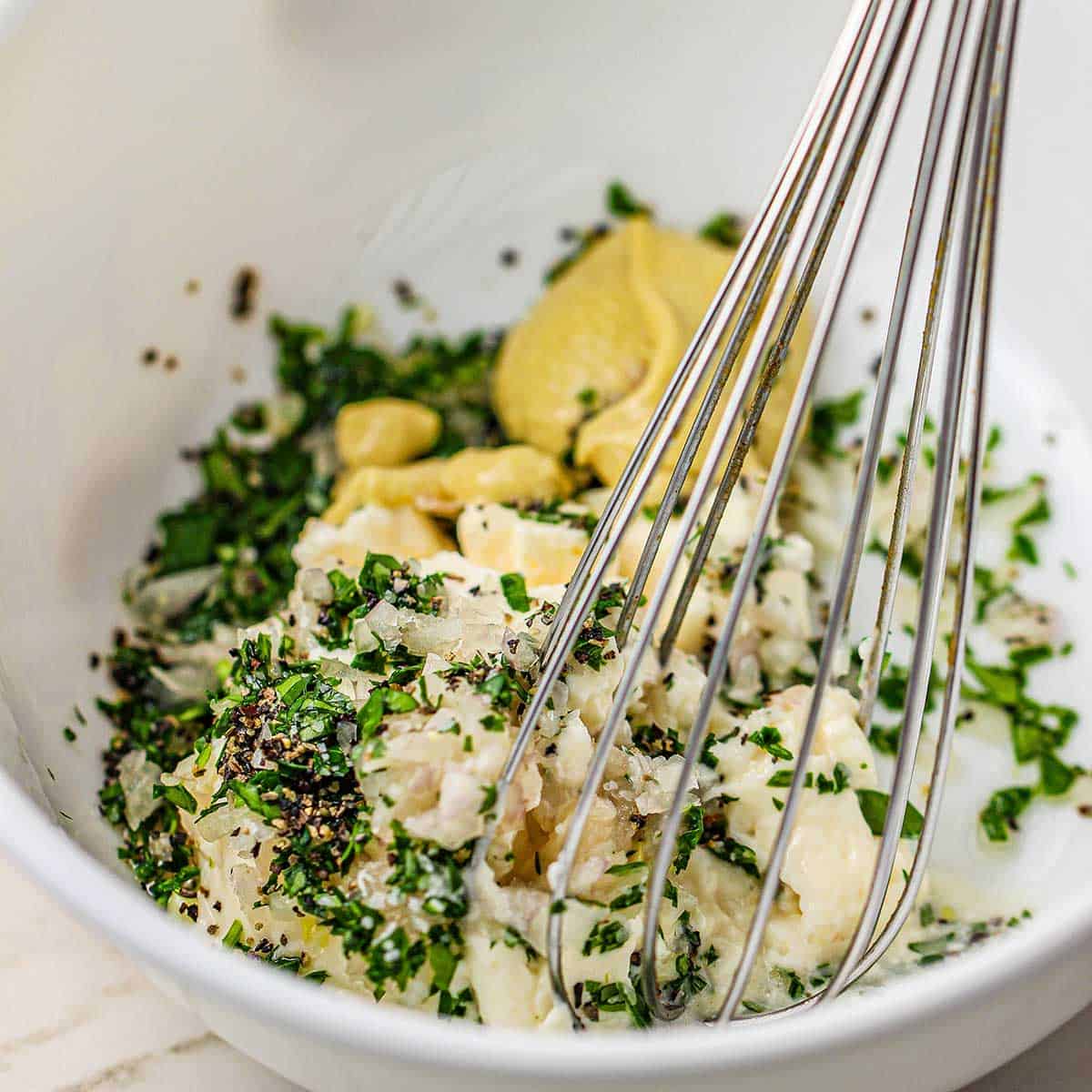 A person using a whisk to combine mayonnaise, Dijon mustard, chopped shallots, salt, and pepper in a small white bowl.