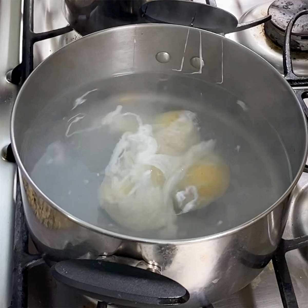 Four eggs being poached in swirling, hot water in a large silver pot on a gas stove.