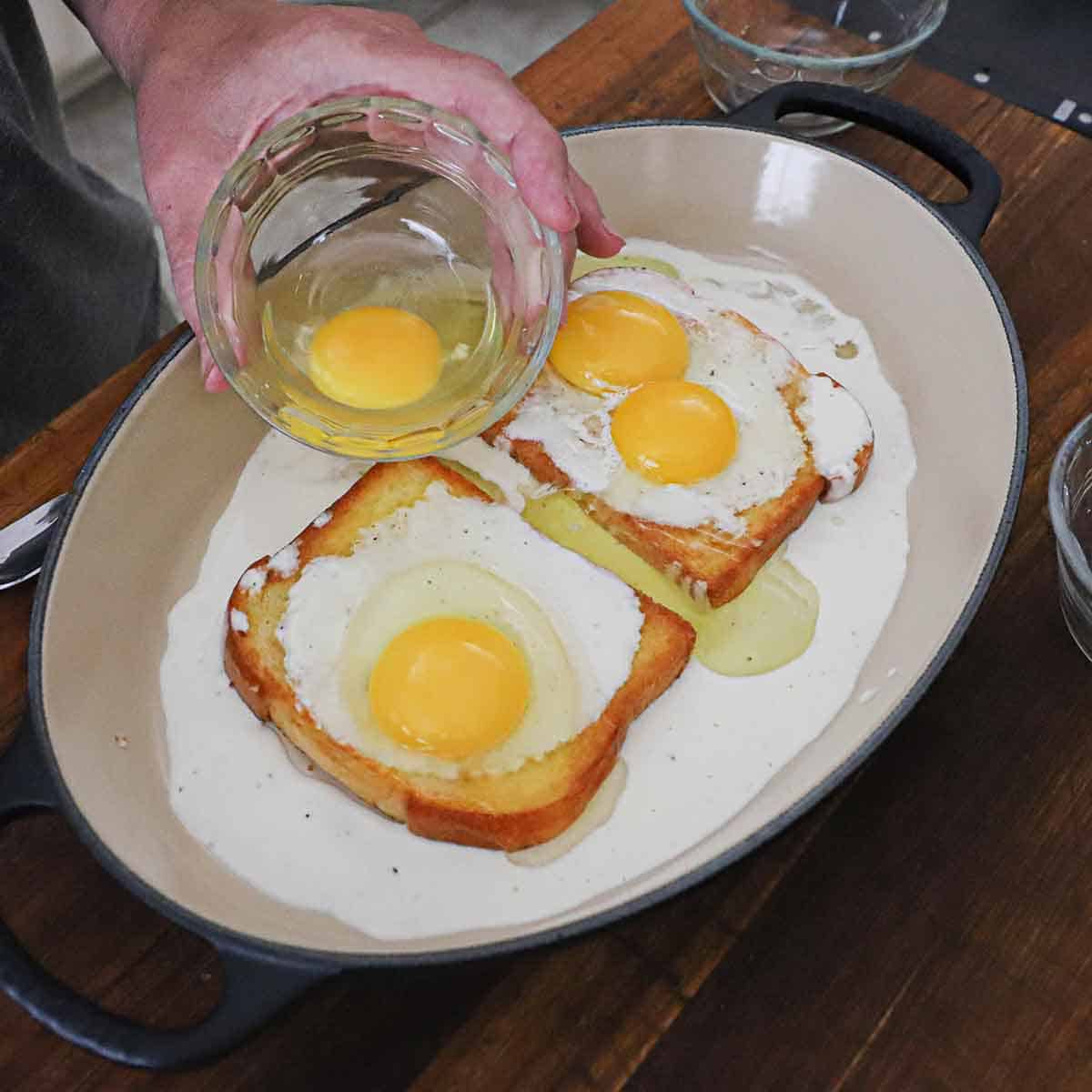 A person holding a small glass bowl with a whole egg in over two slices of bread the resting in a cream sauce with uncooked whole eggs on top of each slice.