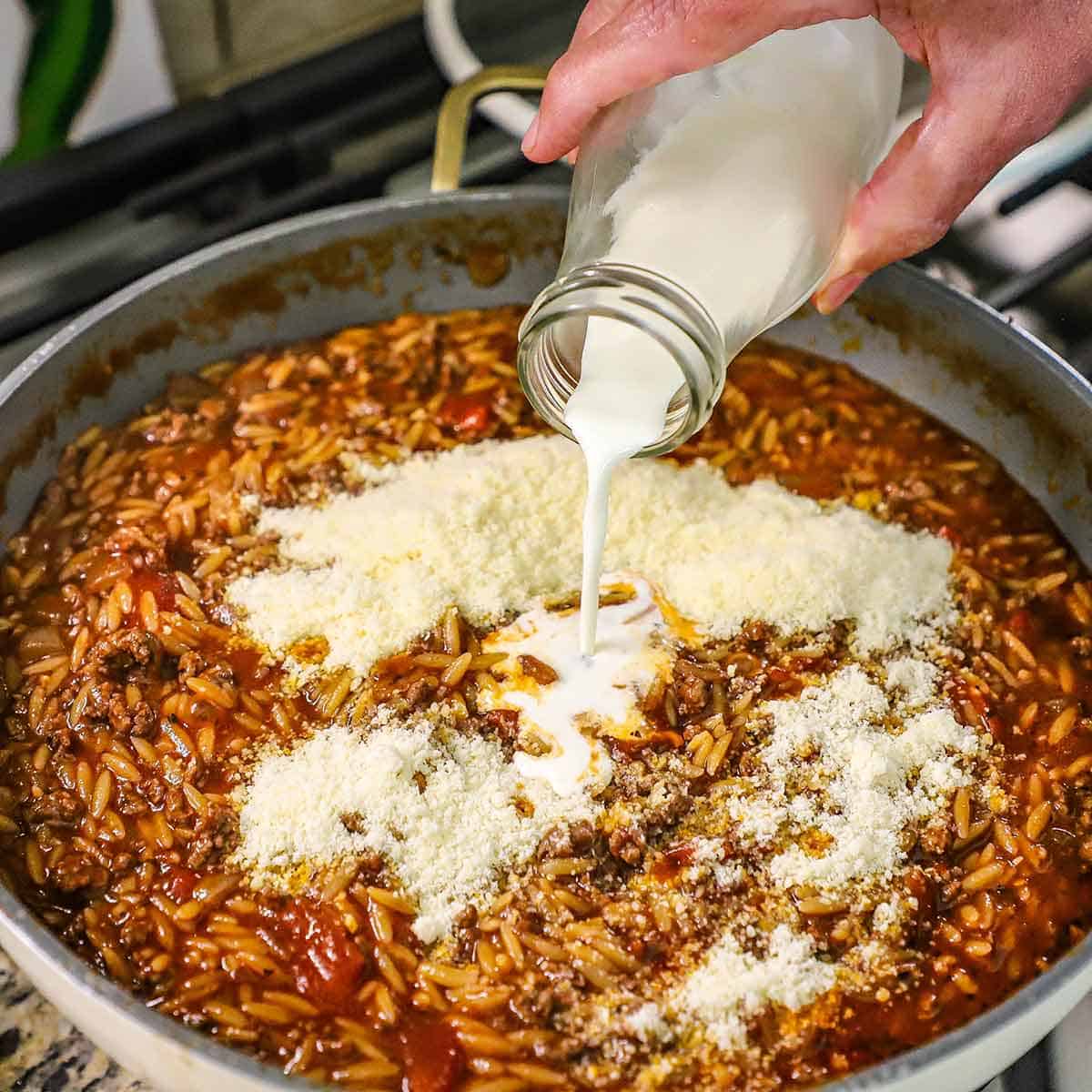 A person pouring heavy cream from a small milk bottle into a pot that is filled with cheesy ground beef orzo and topped with grated Parmesan cheese.