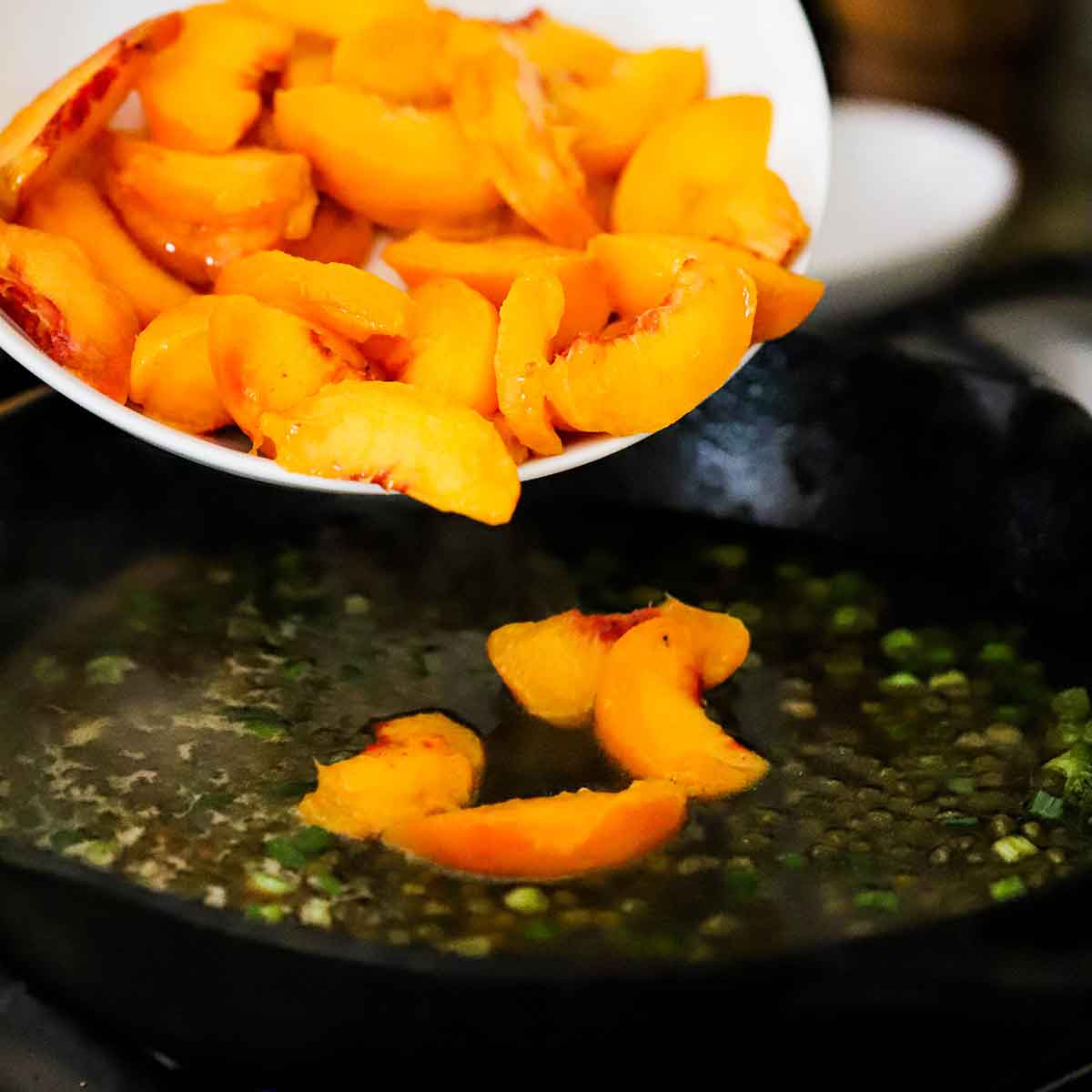 A person transferring slices of fresh peaches into a skillet filled with a simmering wine sauce with capers and chopped scallions.