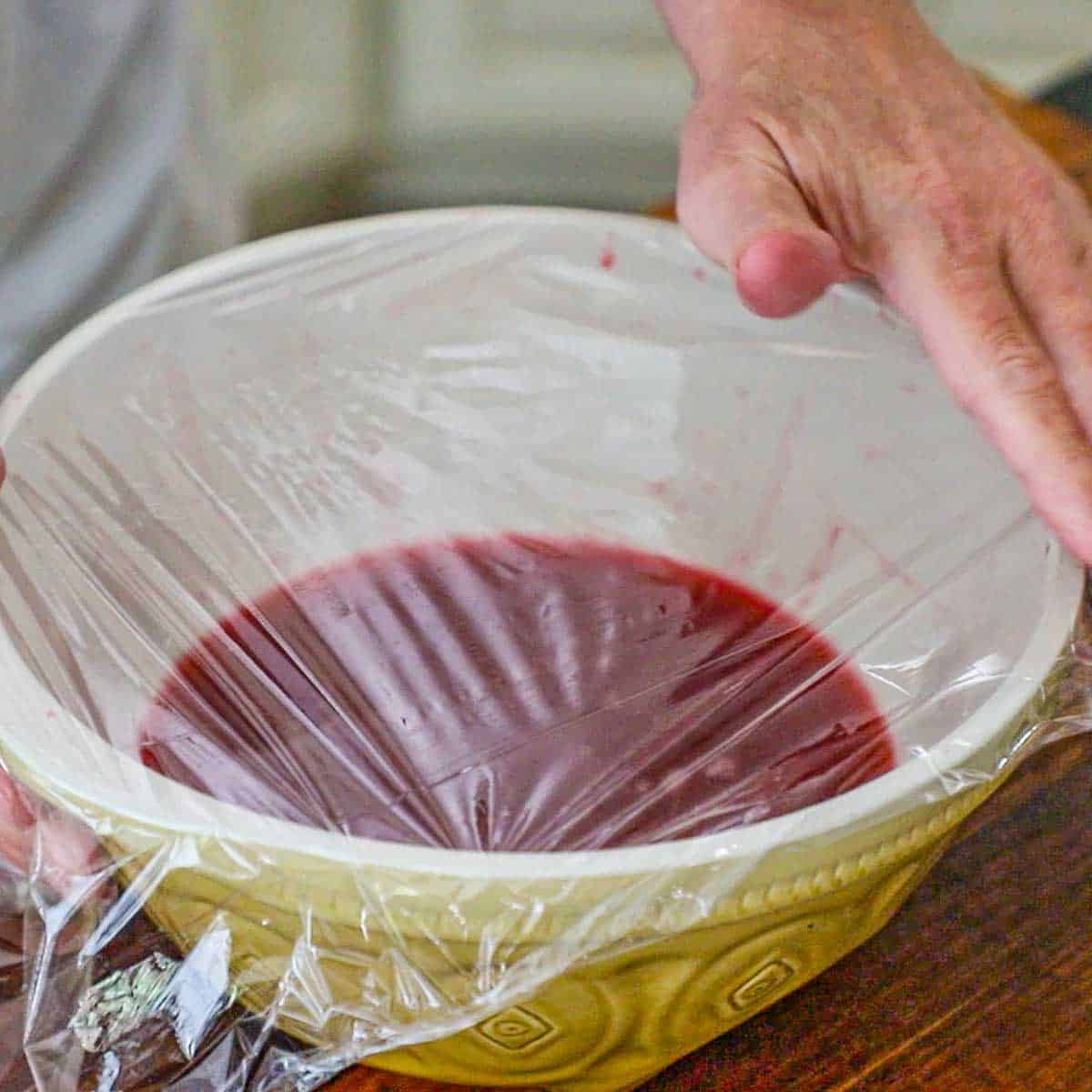 A person covering a ceramic bowl partially filled with a fresh raspberry sorbet mixture with plastic wrap.