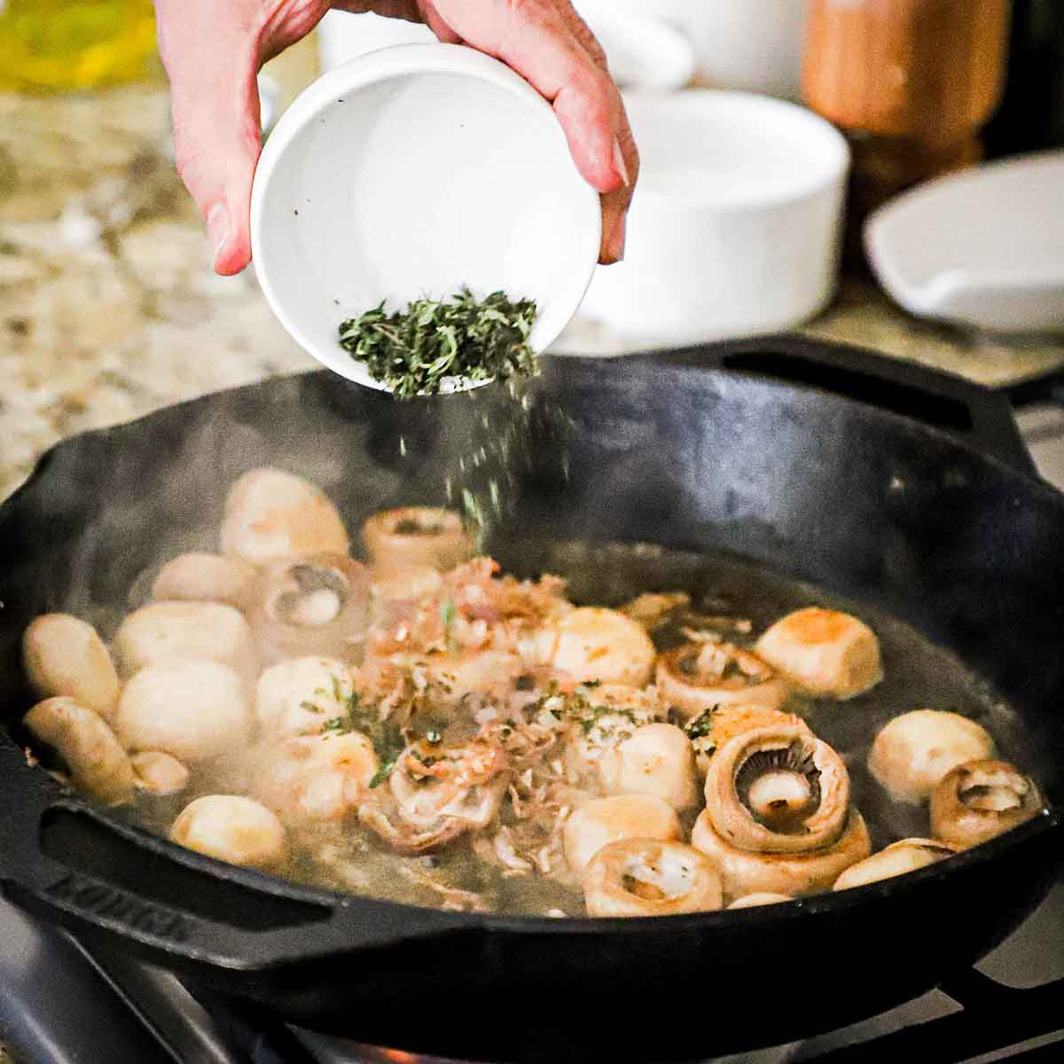 A person transferring chopped fresh thyme from a small white bowl into a cast-iron skillet that is filled with whole sautéed mushrooms.