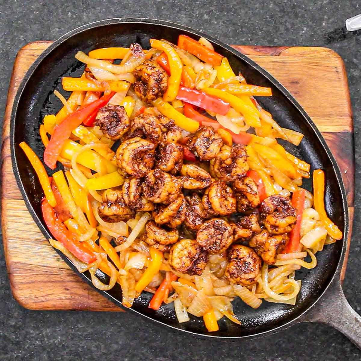 An overhead view of an oversized fajita pan filled with sizzling strips of sautéed onions and bell peppers which are topped with a pile of sautéed seasoned shrimp.