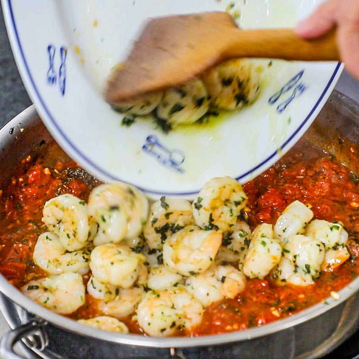 A person using a wooden spatula to transfer sautéed shrimp from a platter into a skillet filled with a simmering marinara sauce.