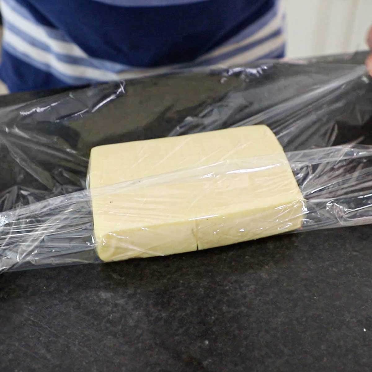 A person wrapping a smoked block of mozzarella in plastic wrap on a black marble counter.