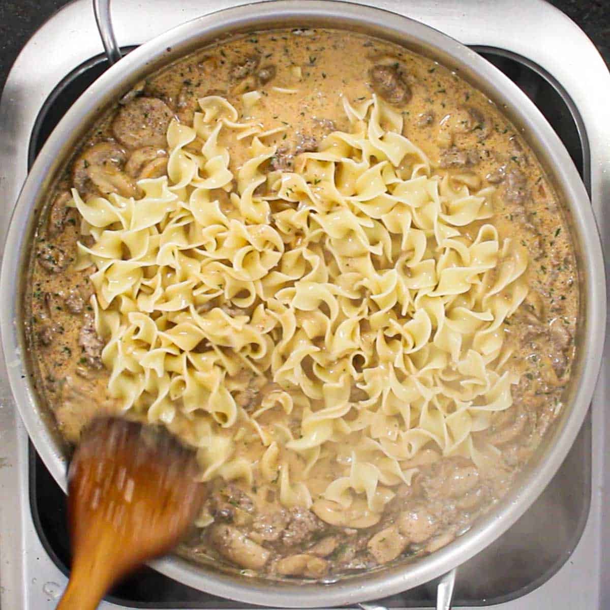 A person using a large wooden spatula to stir cooked egg noodles into a large skillet filled with ground beef stroganoff.
