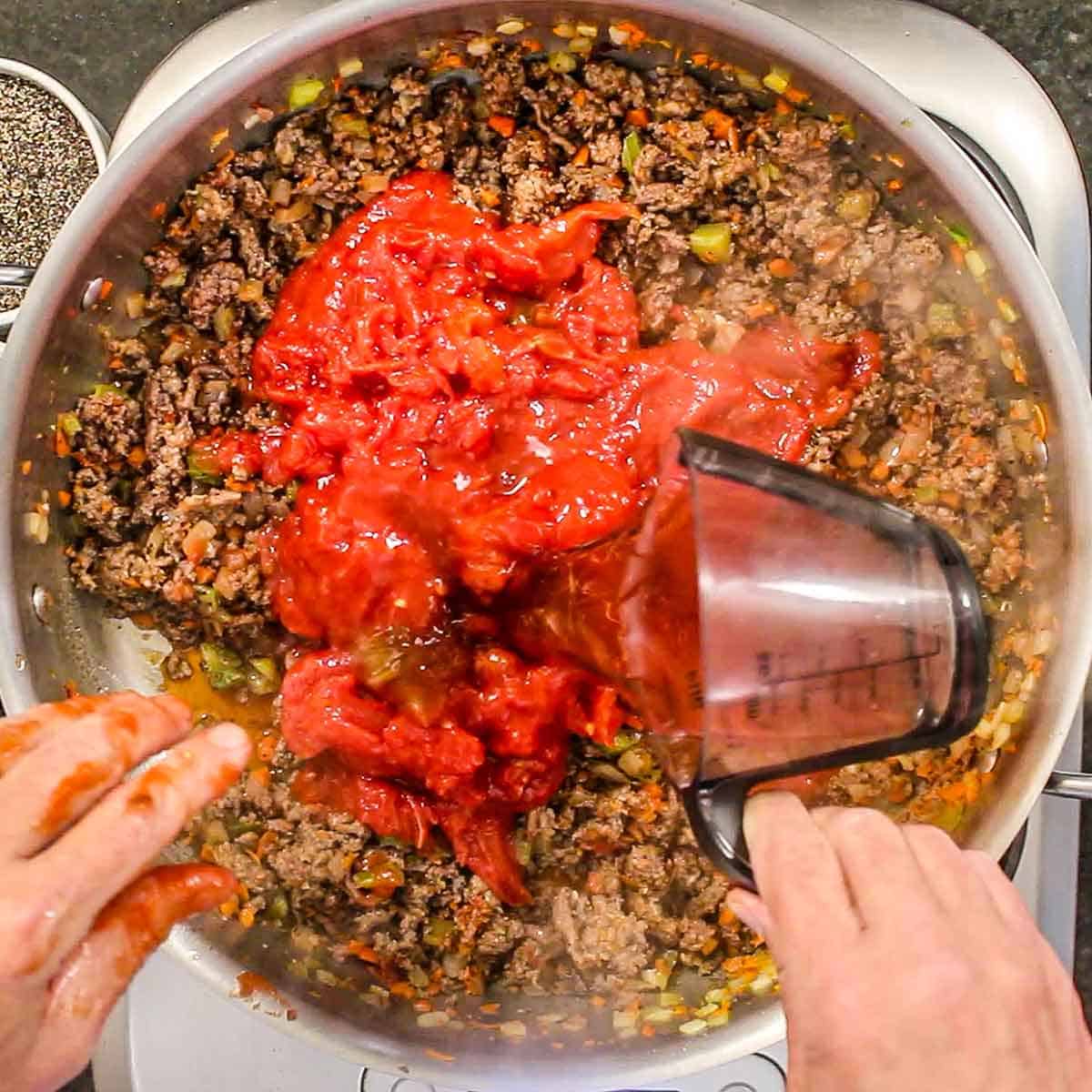A person pouring beef broth from a plastic measuring cup into a skillet filled with a simmering bolognese sauce.