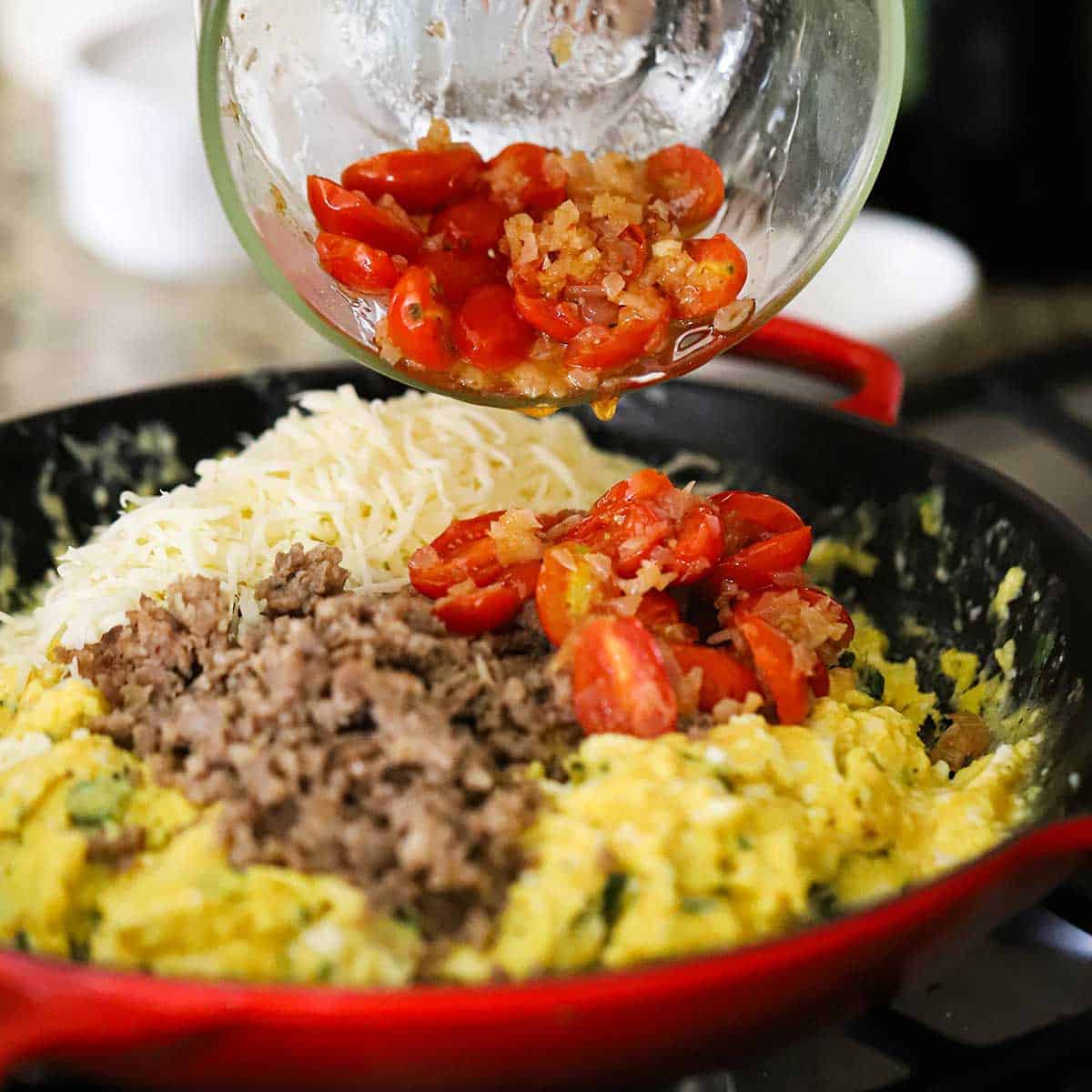 A person adding sautéed cherry tomatoes with shallots into a large skillet holding scrambled eggs, cooked Italian sausage, shredded mozzarella, and fresh basil.