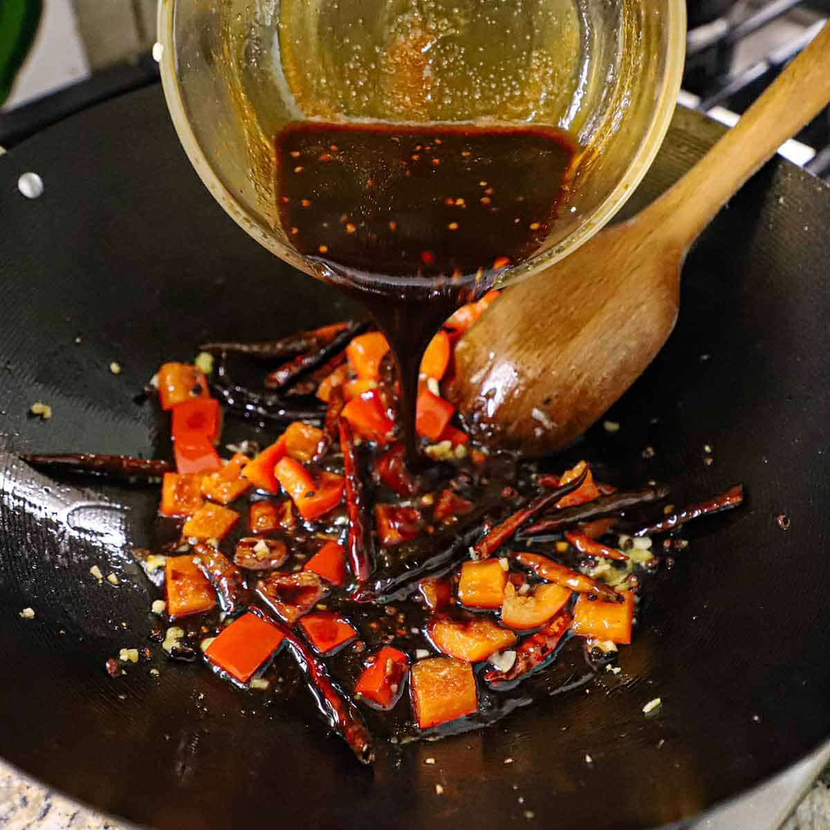 A person pouring a dark brown sticky sauce into a wok that is filled with stir-fried peppers, garlic, and ginger.