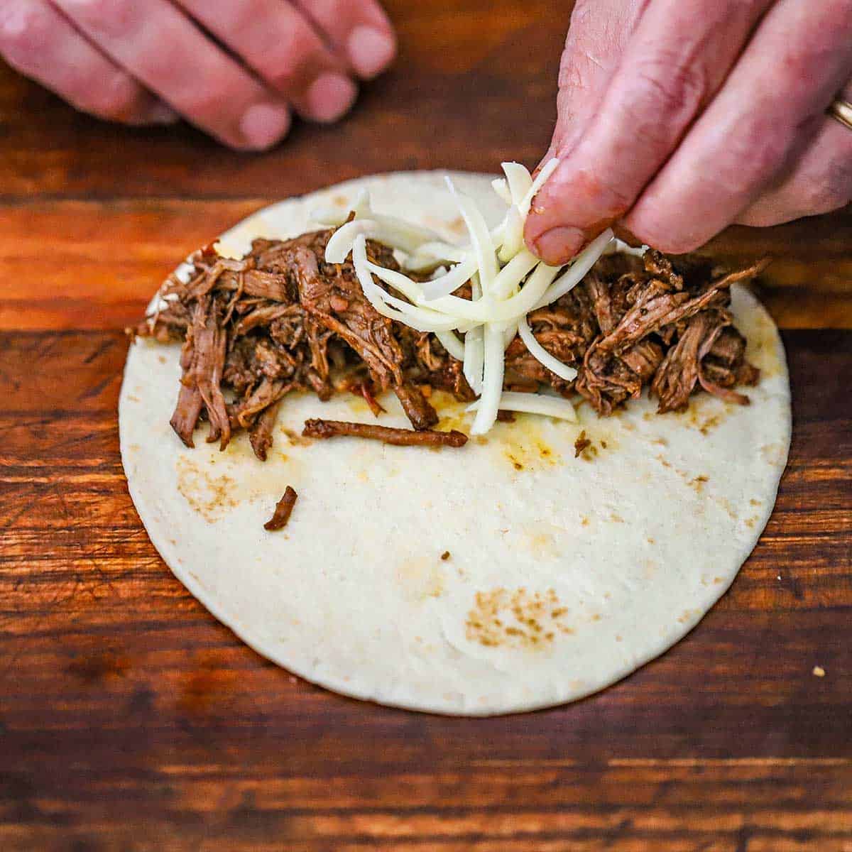 A person placing shredded Monterey Jack cheese over the top of shredded beef on one side of a flour tortilla on a wooden cutting board.