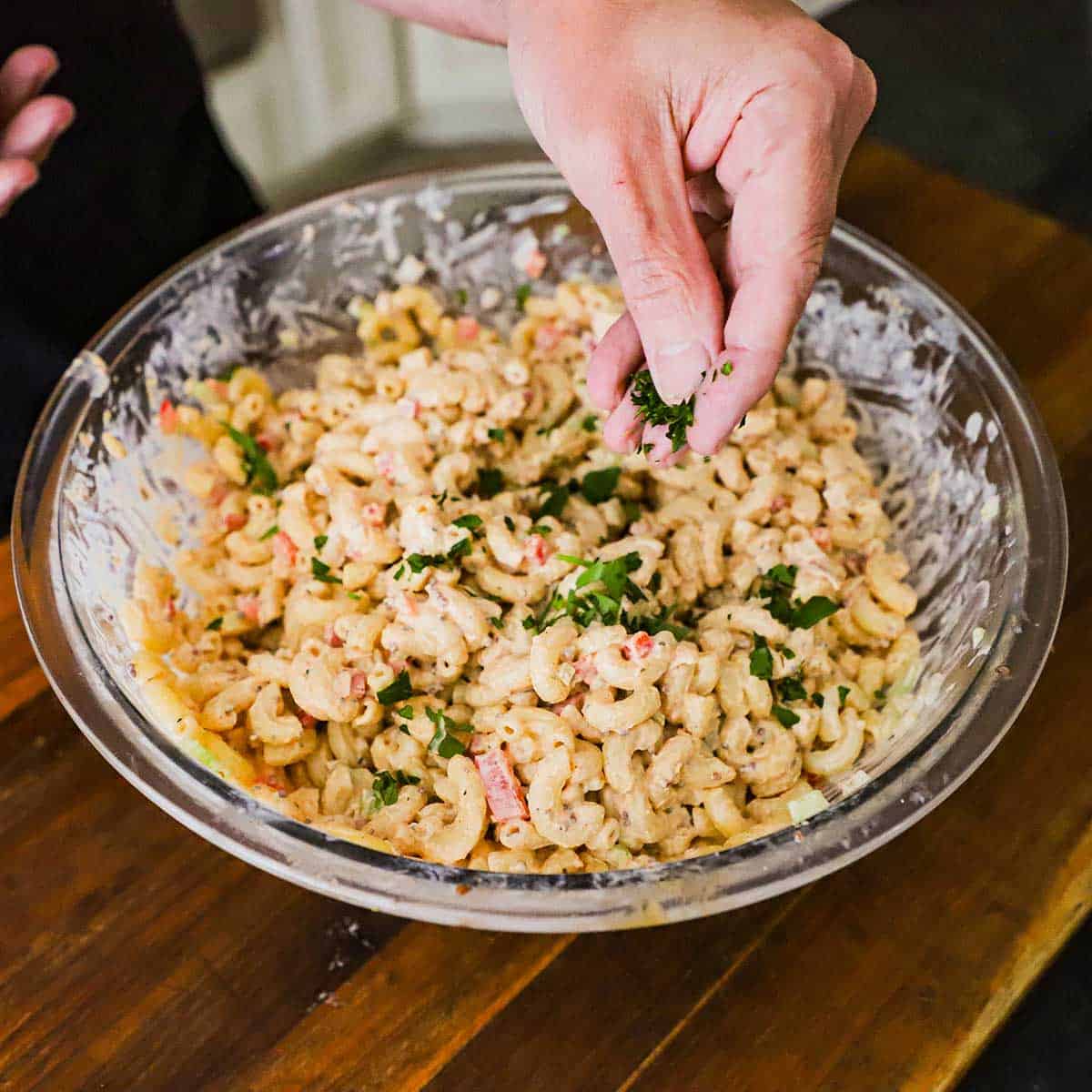 A person sprinkling chopped Italian parsley over the top of a Cajun Pasta Salad that has just been tossed in a large glass bowl.