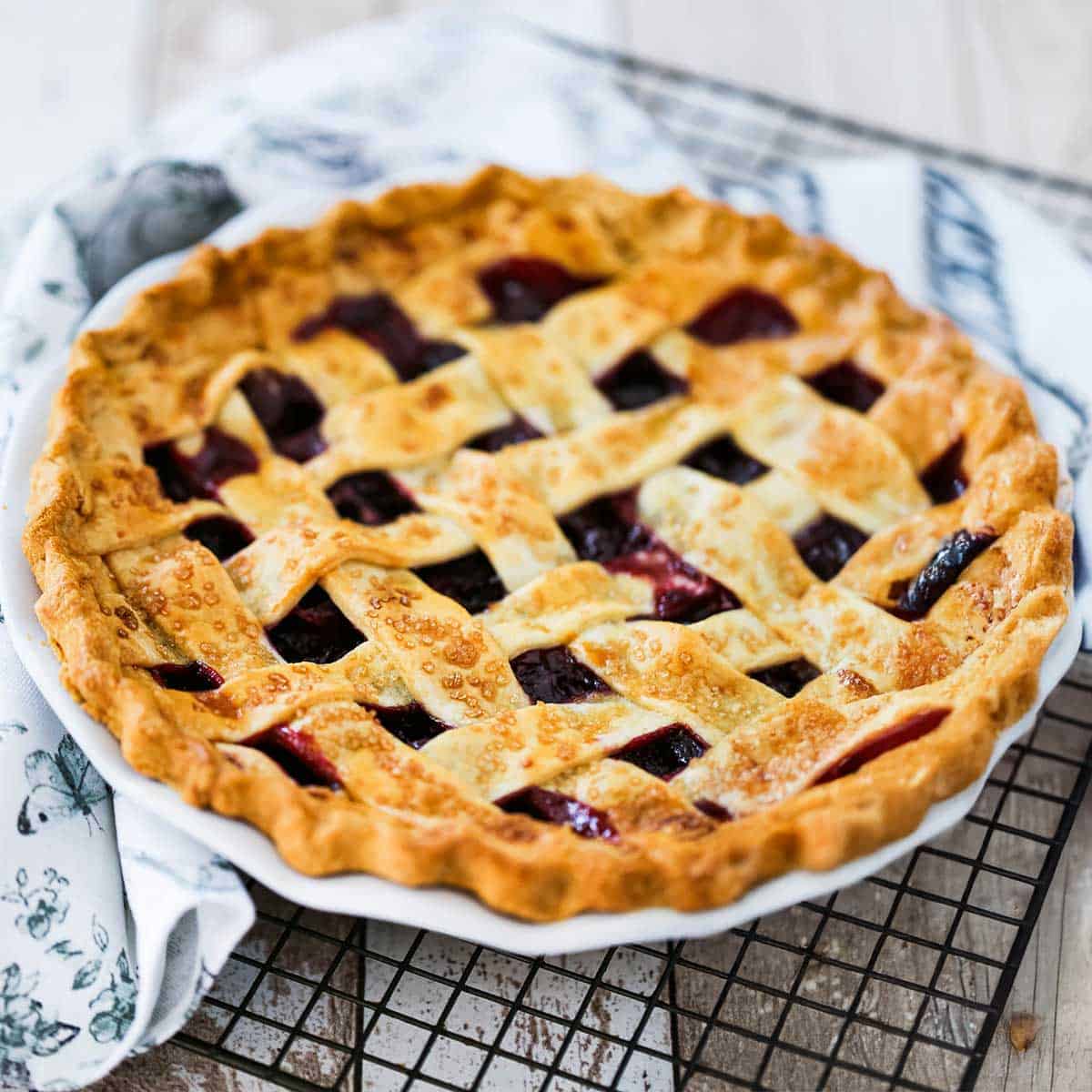 A freshly baked homemade cherry pie topped with a lattice crust resting on a baking rack with a kitchen cloth napkin nearby.