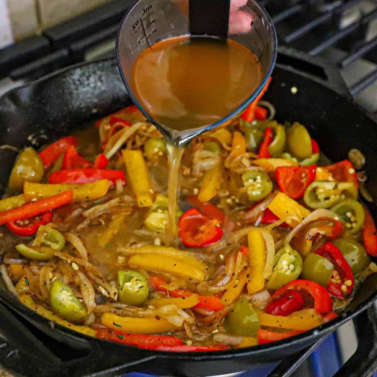A person pouring chicken stock from a small carafe into a large cast-iron skillet filled with simmering sautéed onions, strips of bell peppers, and chopped pickled cherry peppers.