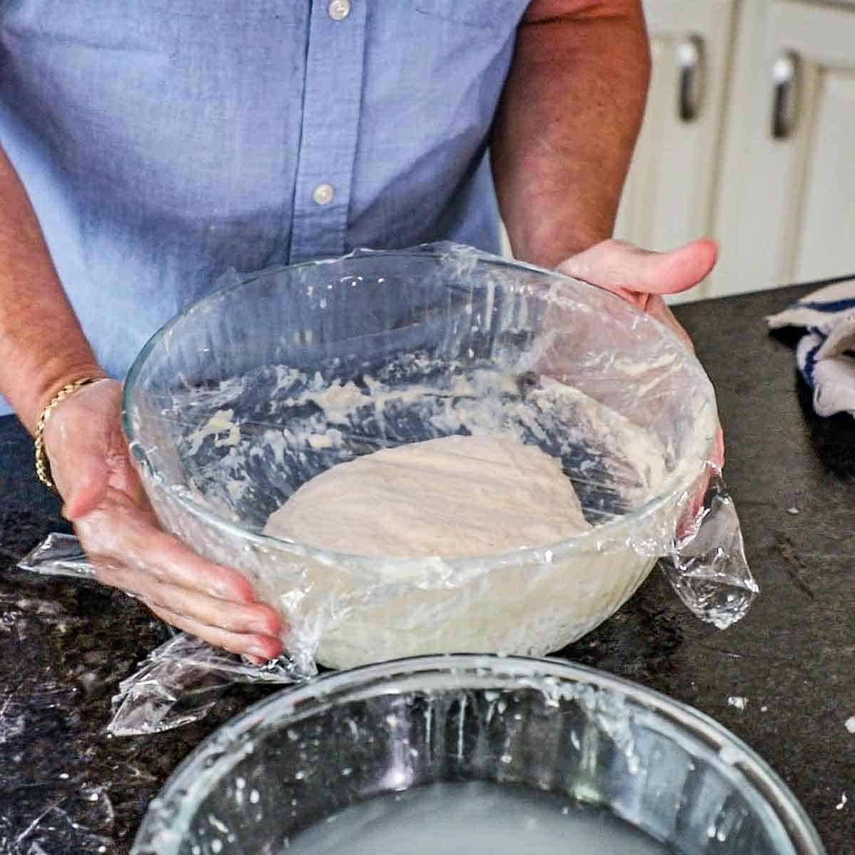 A person placing a large glass bowl that is filled with fermented ciabatta dough with plastic wrap cover the top of the bowl onto a counter.