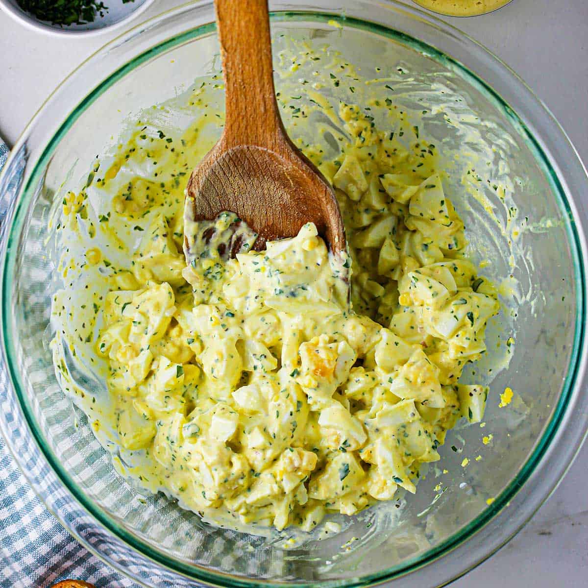 An overhead view of a large glass bowl filled with homemade egg salad with a wooden spoon in the middle of bowl.