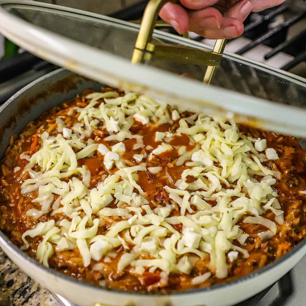 A person placing a glass lid over the top of a large skillet filled with hot ground beef orzo that has been topped with shredded mozzarella cheese.