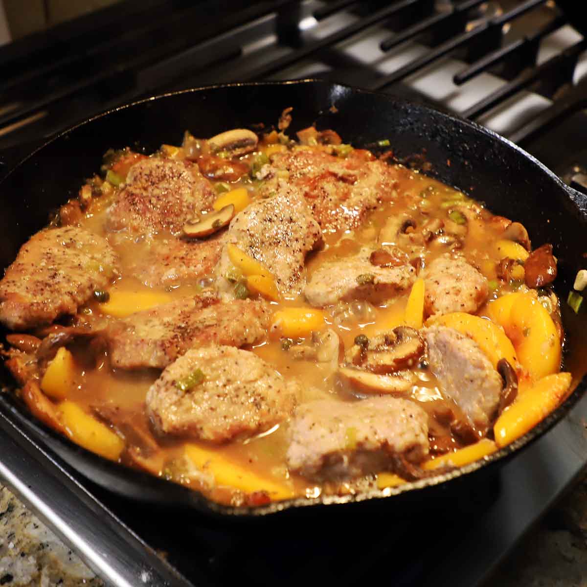 A large black cast-iron skillet filled with pan-fried pork cutlets with peaches simmering in a sauce with capers and scallions.