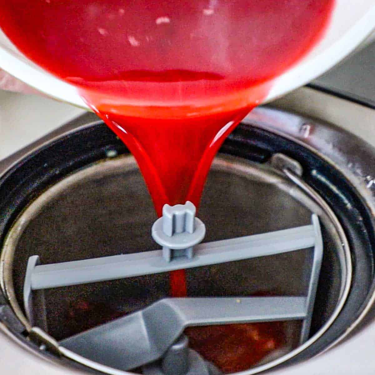 A person pouring a fresh raspberry sorbet mixture into the bucket of an electric ice cream maker.