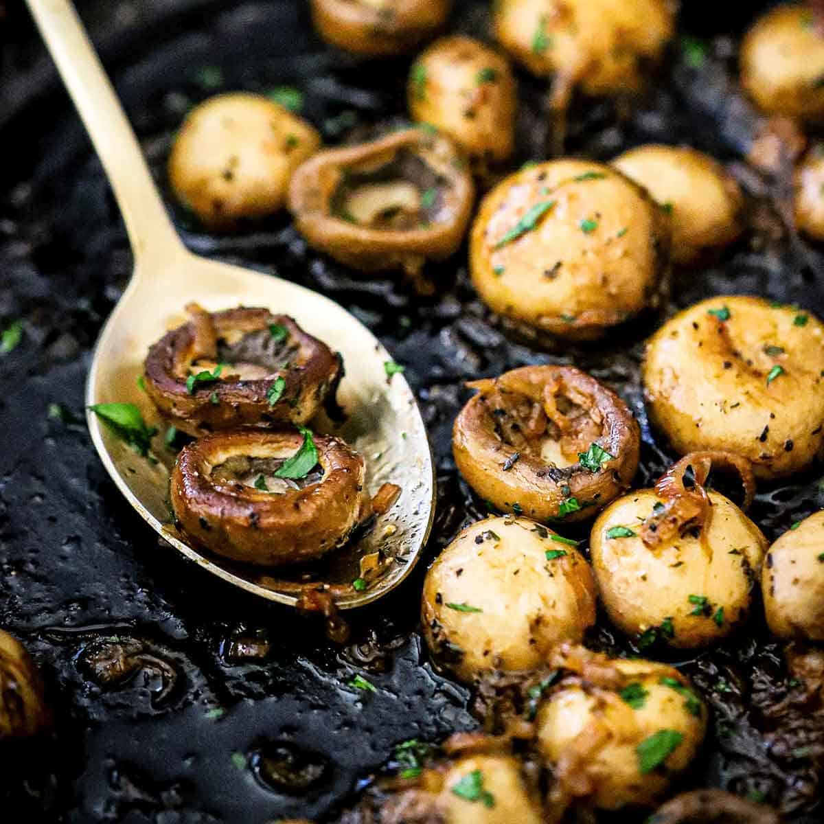 A close-up view of whole sautéed mushrooms resting in a black cast-iron skillet and on a gold serving spoon also resting in the skillet.
