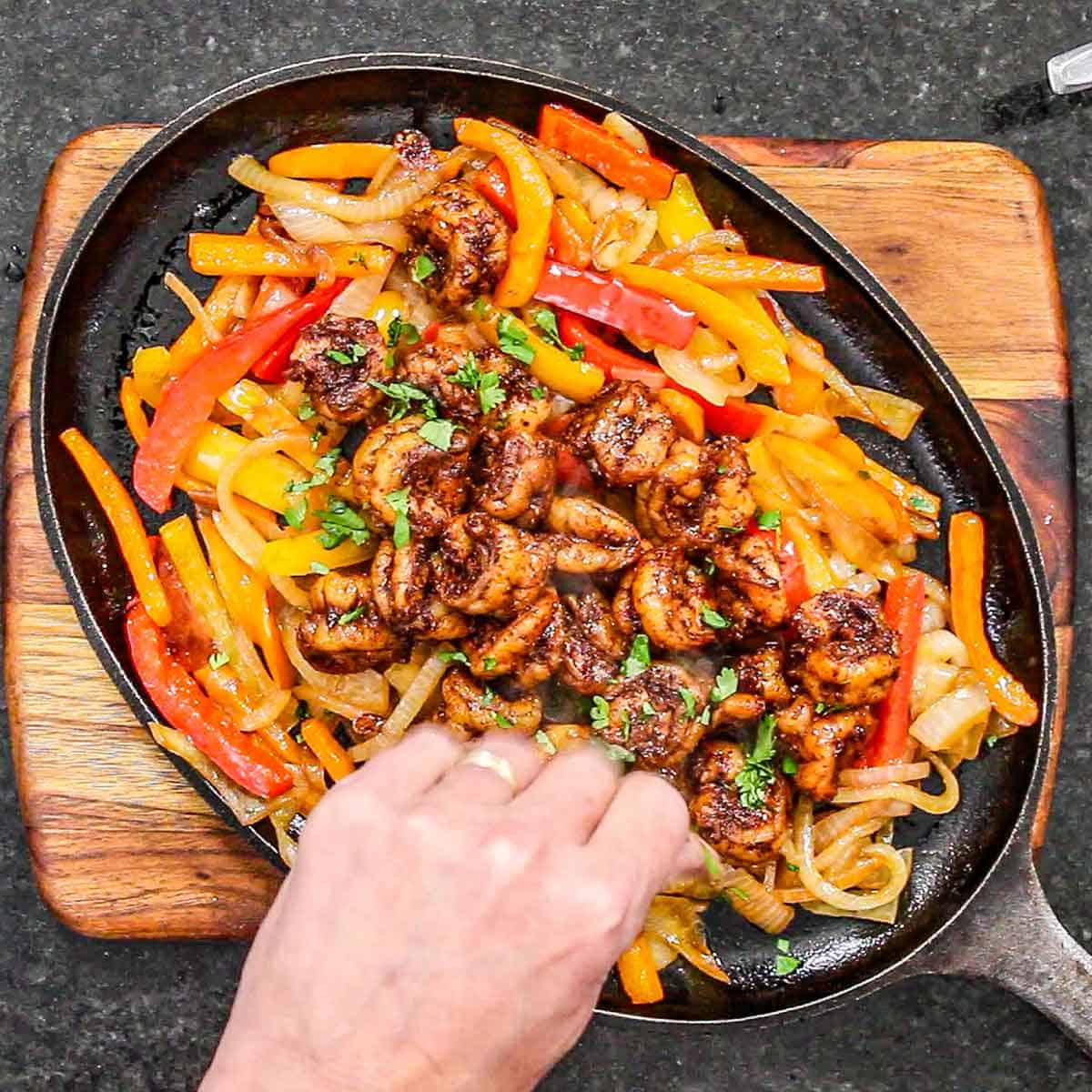 A person sprinkling chopped cilantro over the top of a large fajita skillet filled with a large serving of best shrimp fajitas.