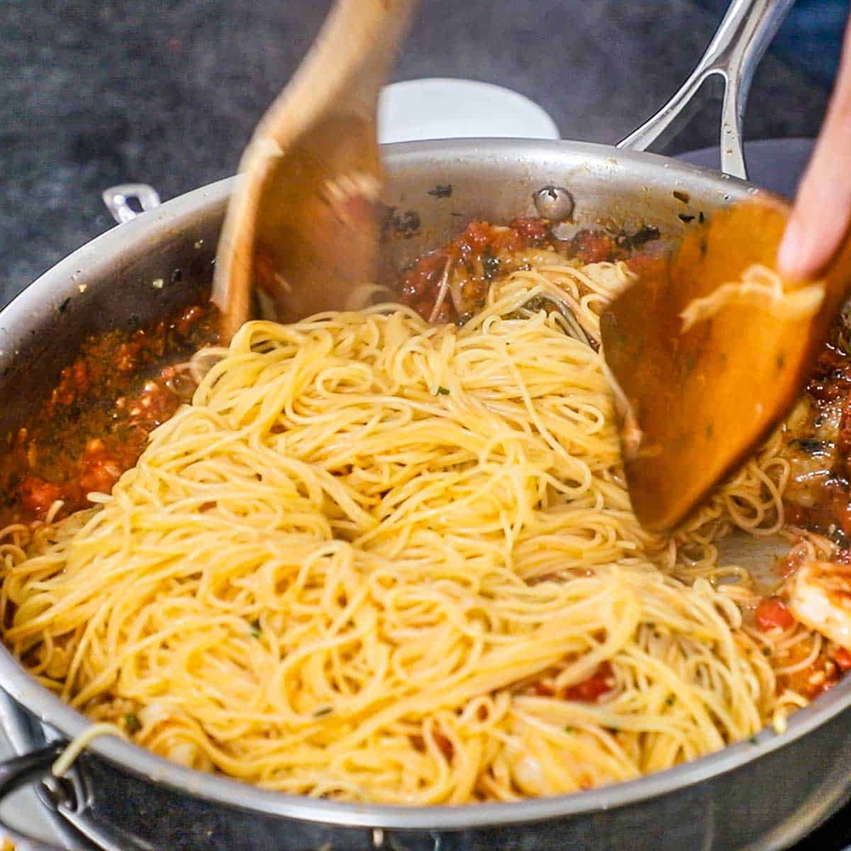 A person using a large wooden spoon and spatula to toss cooked angel hair pasta in a large skillet filled with shrimp marinara.