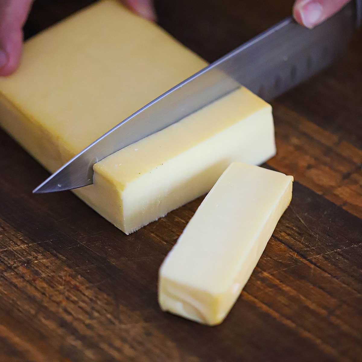 A person using a large chef's knife to slice into a block of smoked mozzarella on a wooden cutting board.