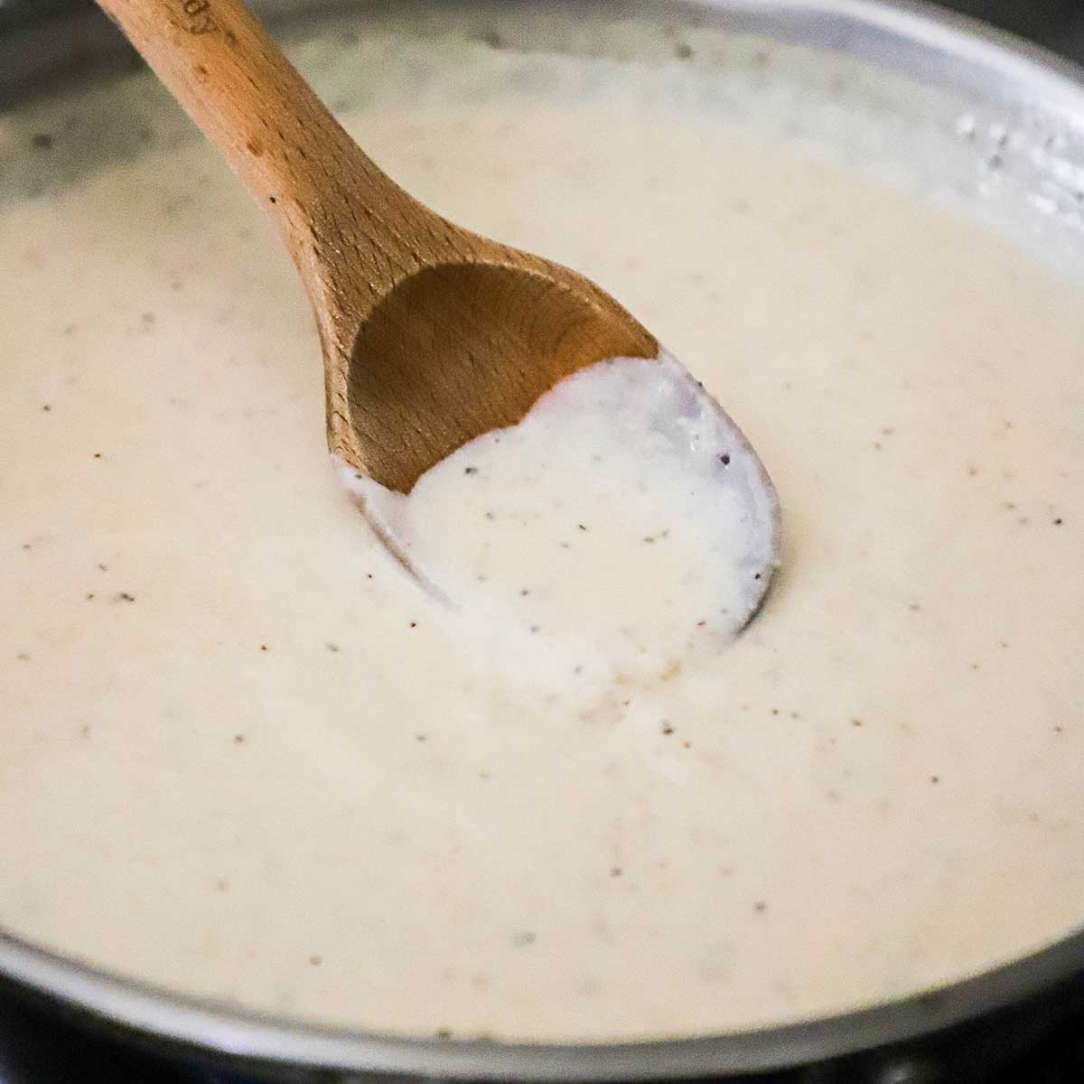 A close-up view of a wooden spoon being used to stir cream gravy in a large silver skillet.