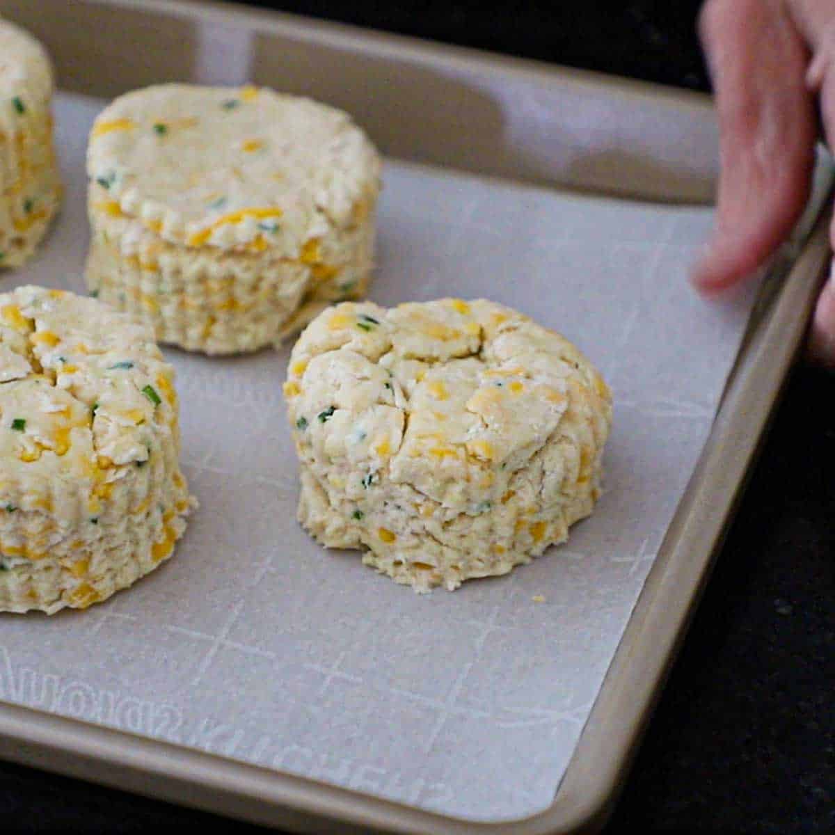 A person holding a baking pan lined with parchment paper and topped with uncooked flaky cheddar biscuits.