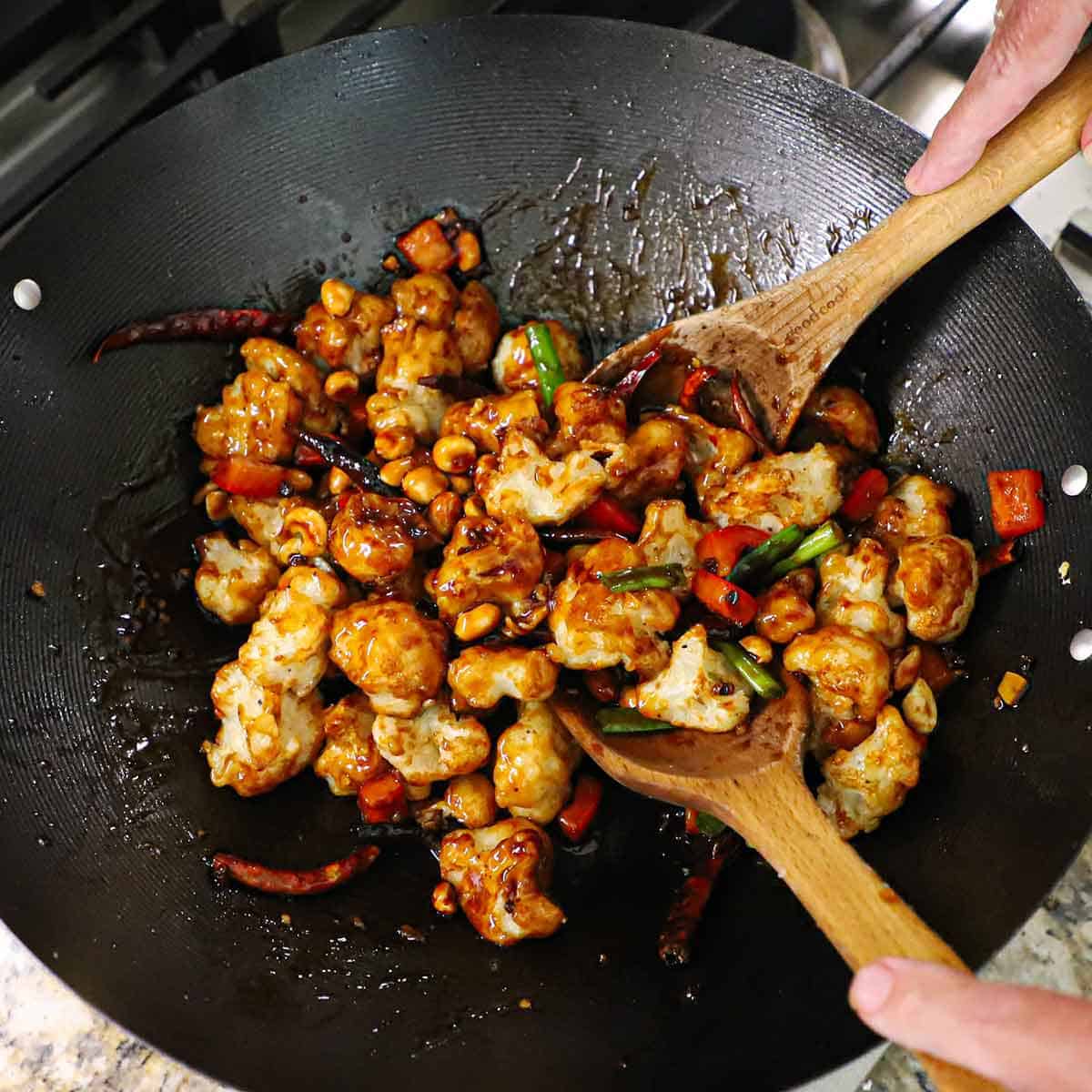 A person using two large wooden spoons to toss deep-fried cauliflower florets in a Chinese kung pao sauce with peppers and peanuts.