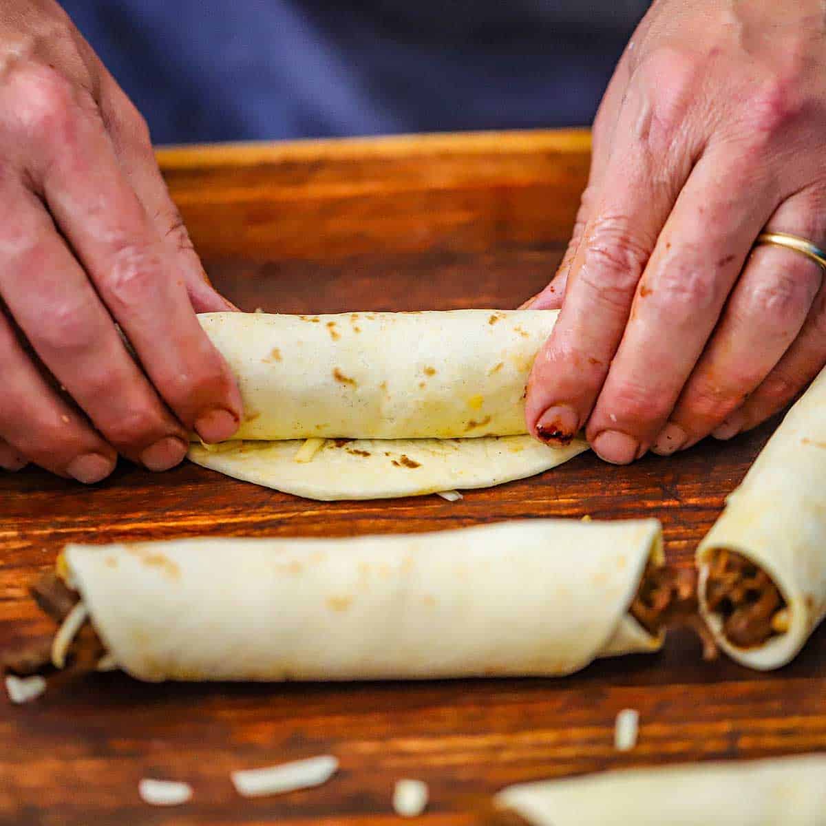 A person using two hands to tightly roll a tortilla that is filled with shredded braised beef and cheese.