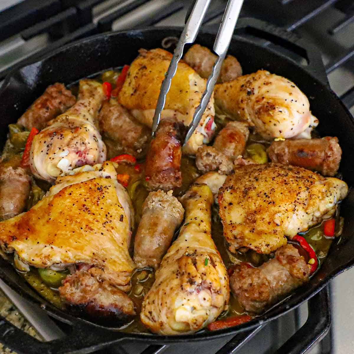 A person using a pair of metal tongs to nestle a seared chunk of Italian sausage into a large cast-iron skillet filled with seared chicken pieces in a scarpariello sauce on a gas stove.