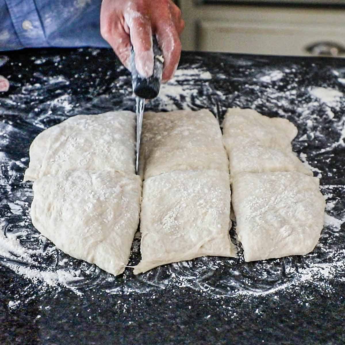 A person using a bench scraper to cut a rectangular piece of dough into squares on a heavily floured marble counter.