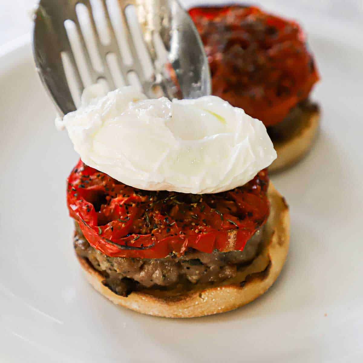 A person using a slotted spoon to place a freshly poached egg on top of a roasted tomato slice that is resting on a cooked breakfast sausage patty on a toasted English muffin.