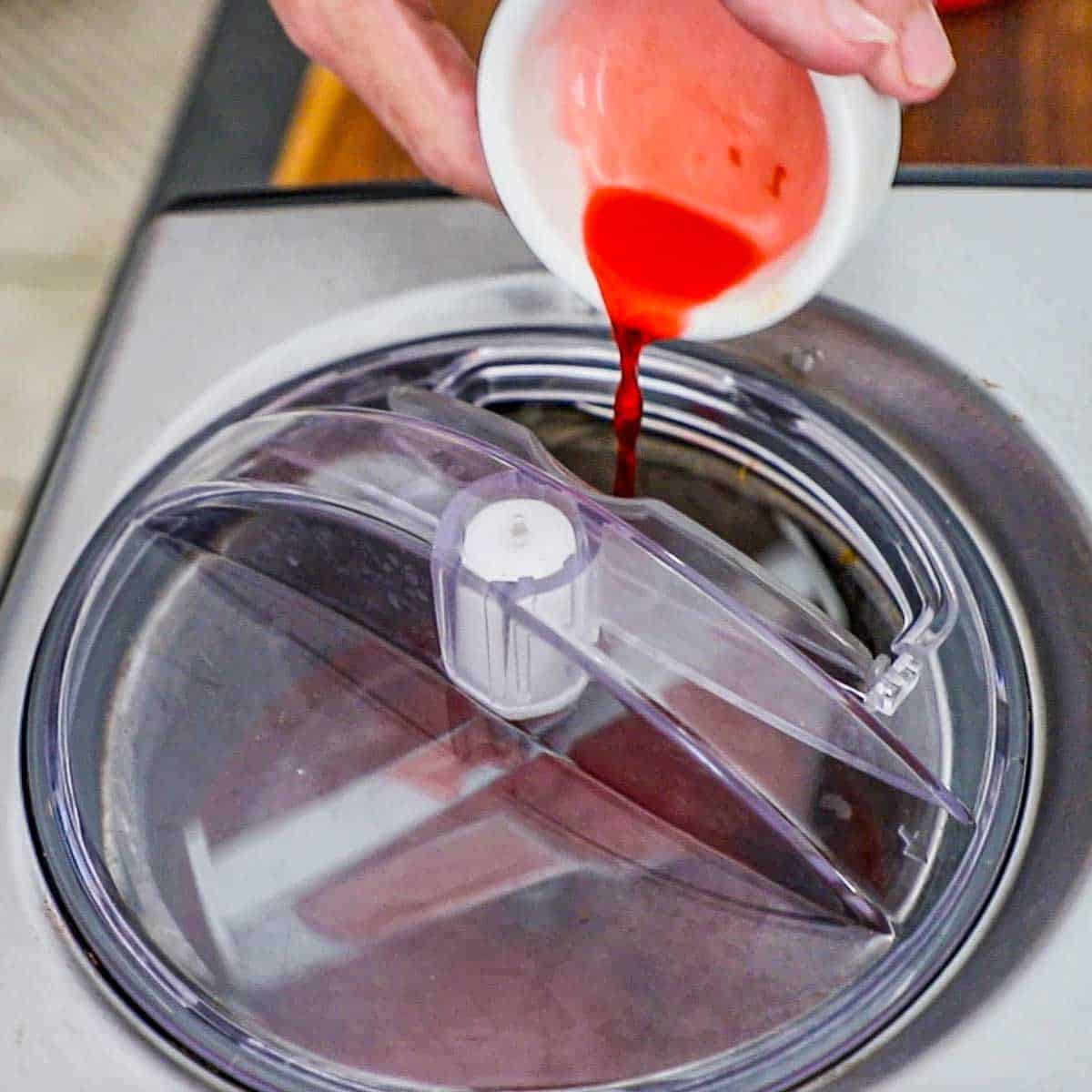 A person pouring a raspberry liqueur from a small white bowl through an opening in a lid on top of a churning electric ice cream maker.