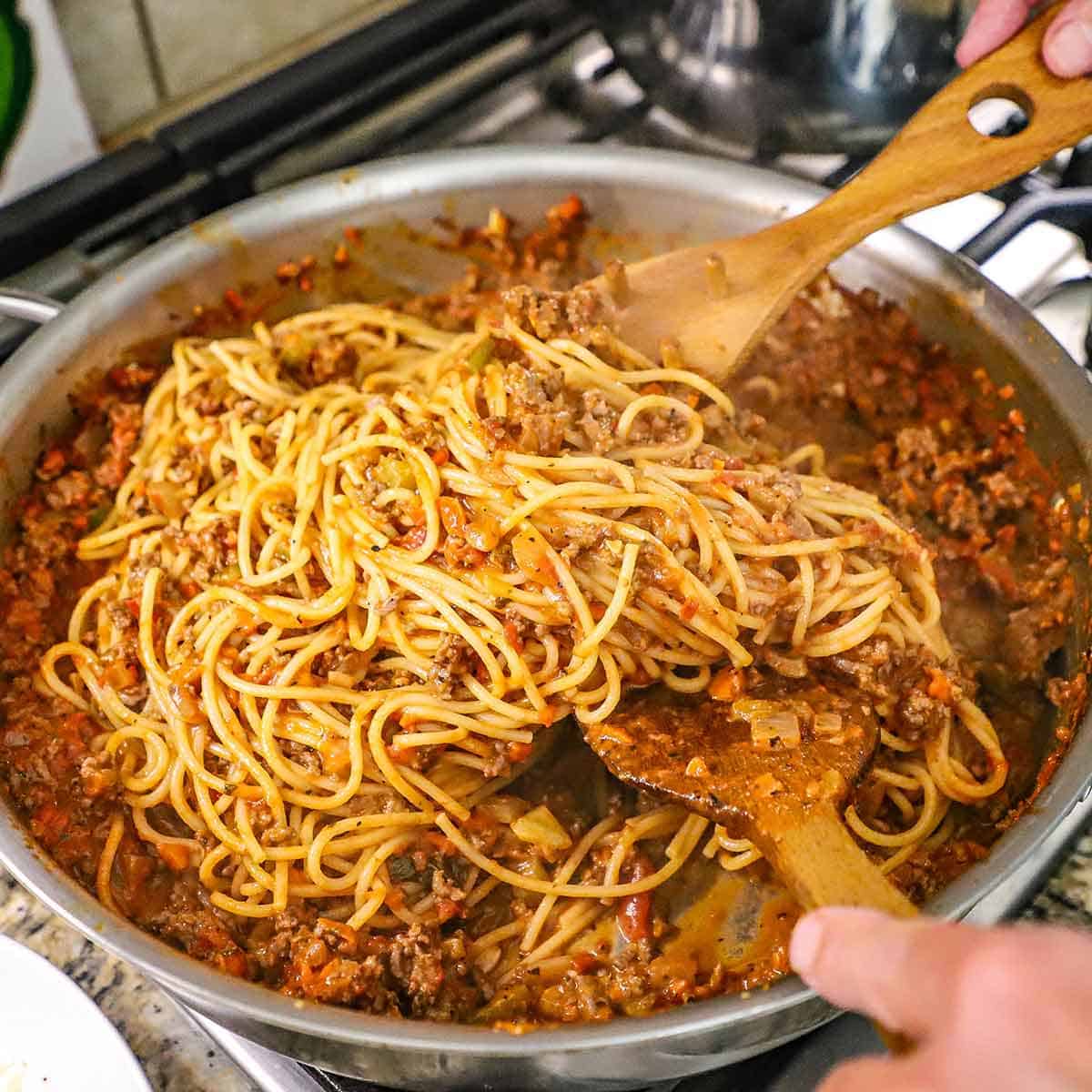 A person using two wooden spatulas to toss cooked spaghetti noodles with a tomato bolognese sauce in a large skillet on a gas stove.