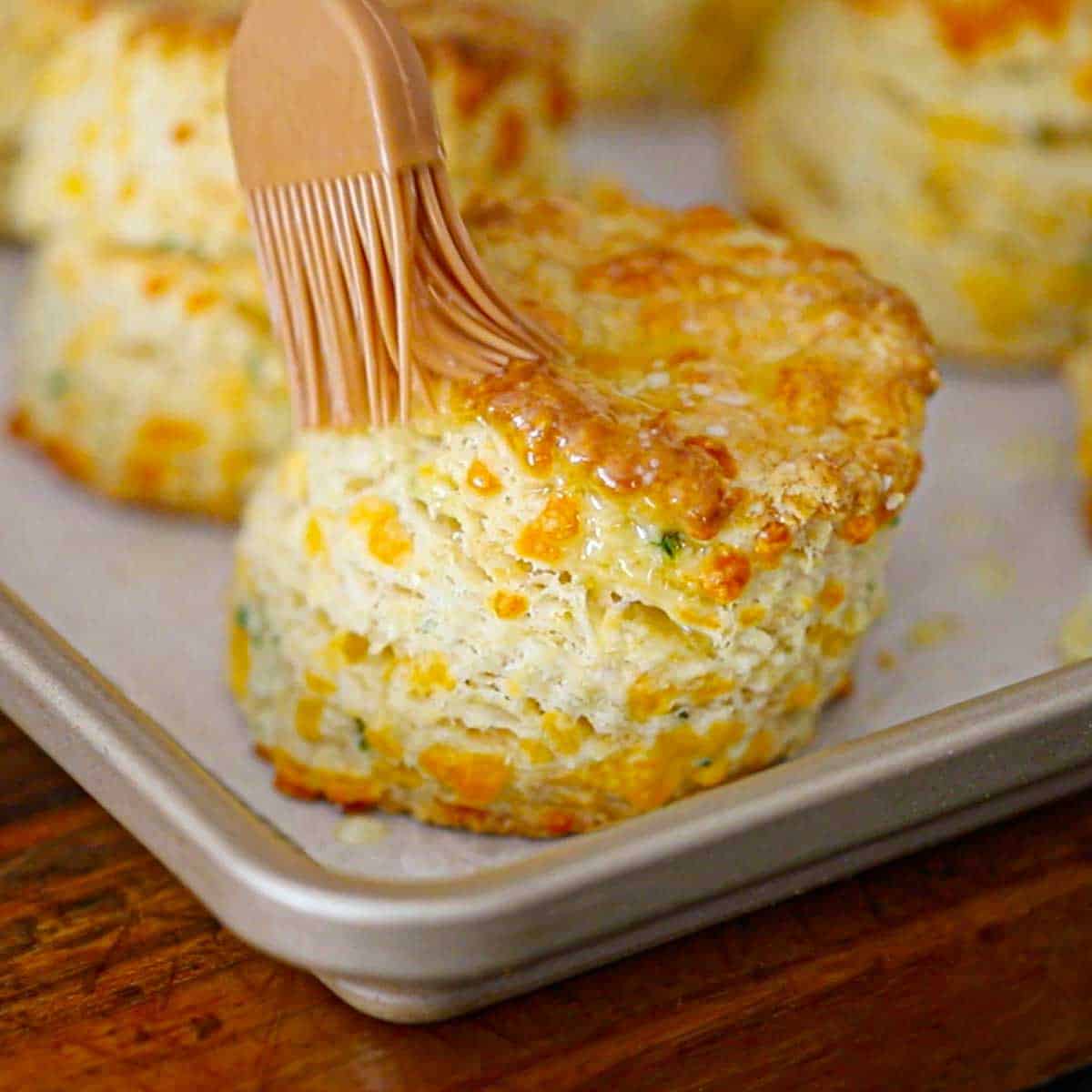 A pastry brush being used to brush melted butter over the tops of freshly baked flaky cheddar biscuits.