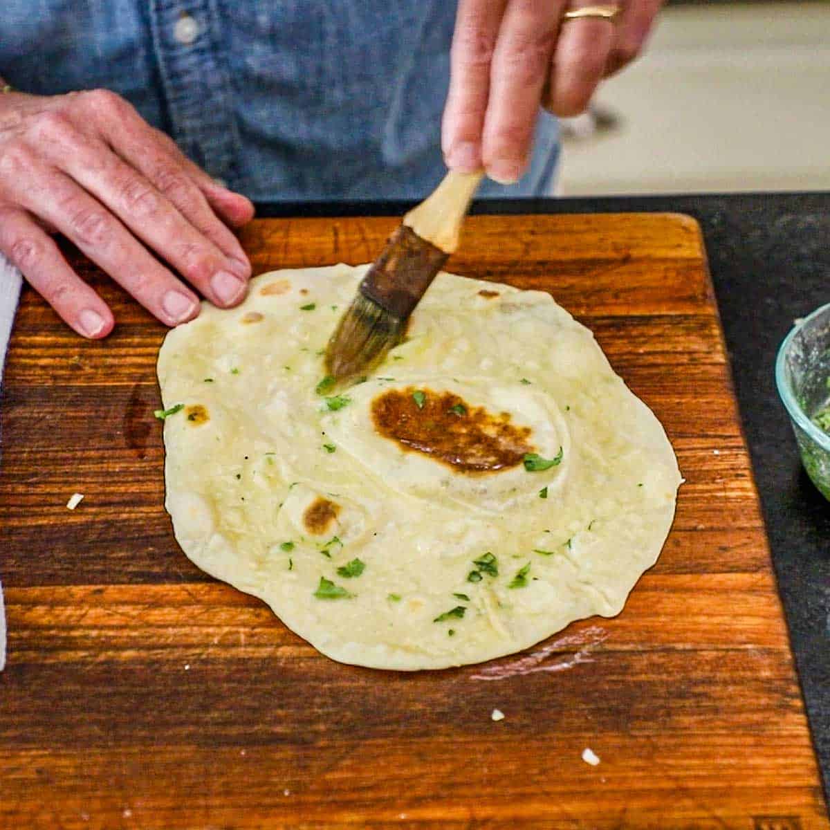 A person using a pastry brush to apply melted garlic-cilantro butter over the surface of a freshly prepared homemade garlic naan on a wooden cutting board.