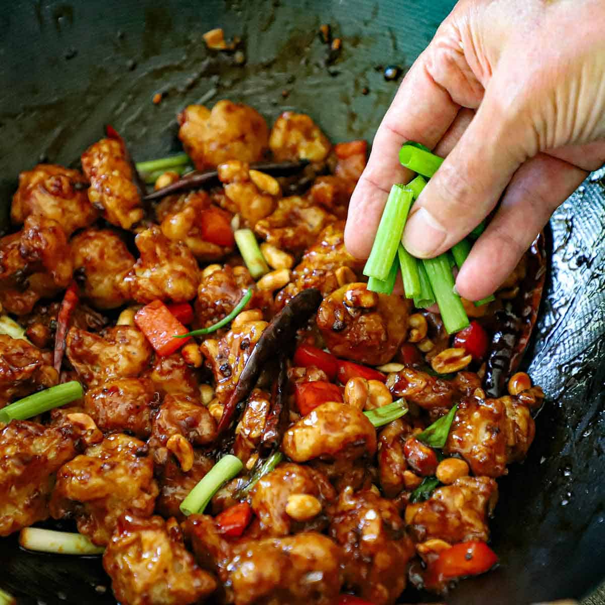 A person using his fingers to sprinkle strips of scallions over the top of a wok filled with a freshly prepared crispy kung pao cauliflower.