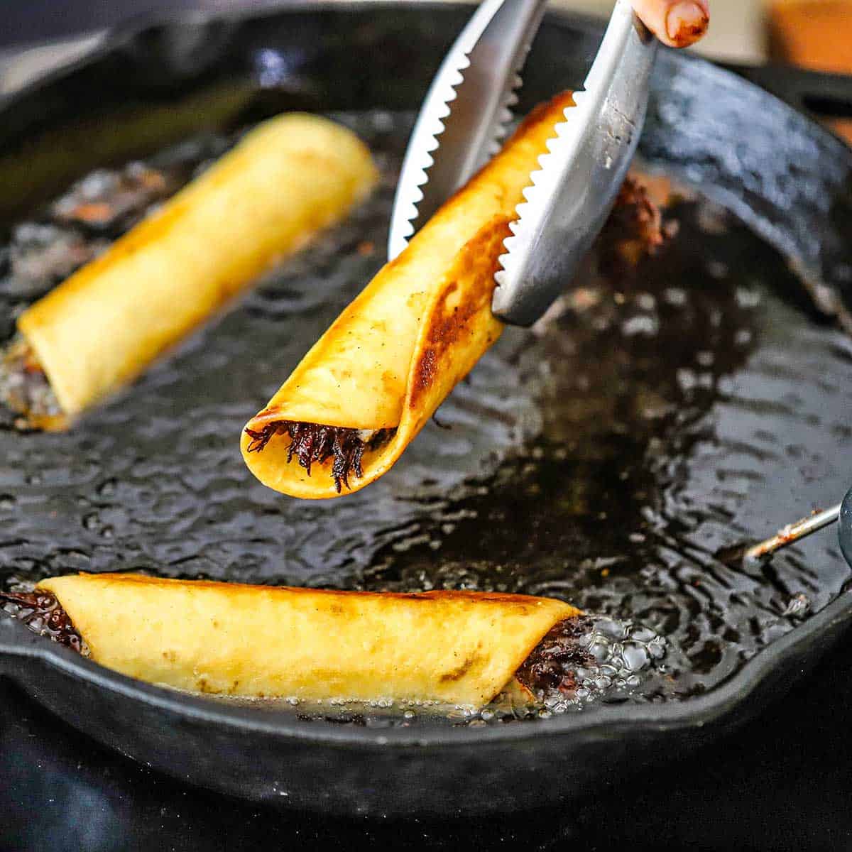 A person using a pair of metal tongs to lift a crispy beef flauta from hot oil in a cast-iron skillet.