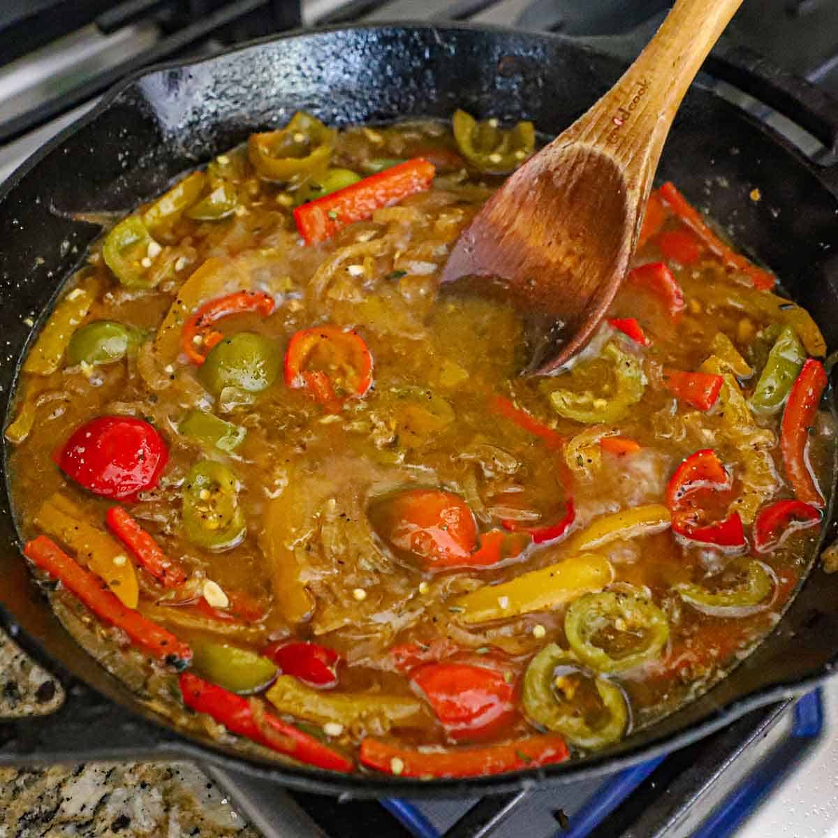 A person using a wooden spoon to stir a simmering scarpariello sauce in a large cast-iron skillet on a gas stove.