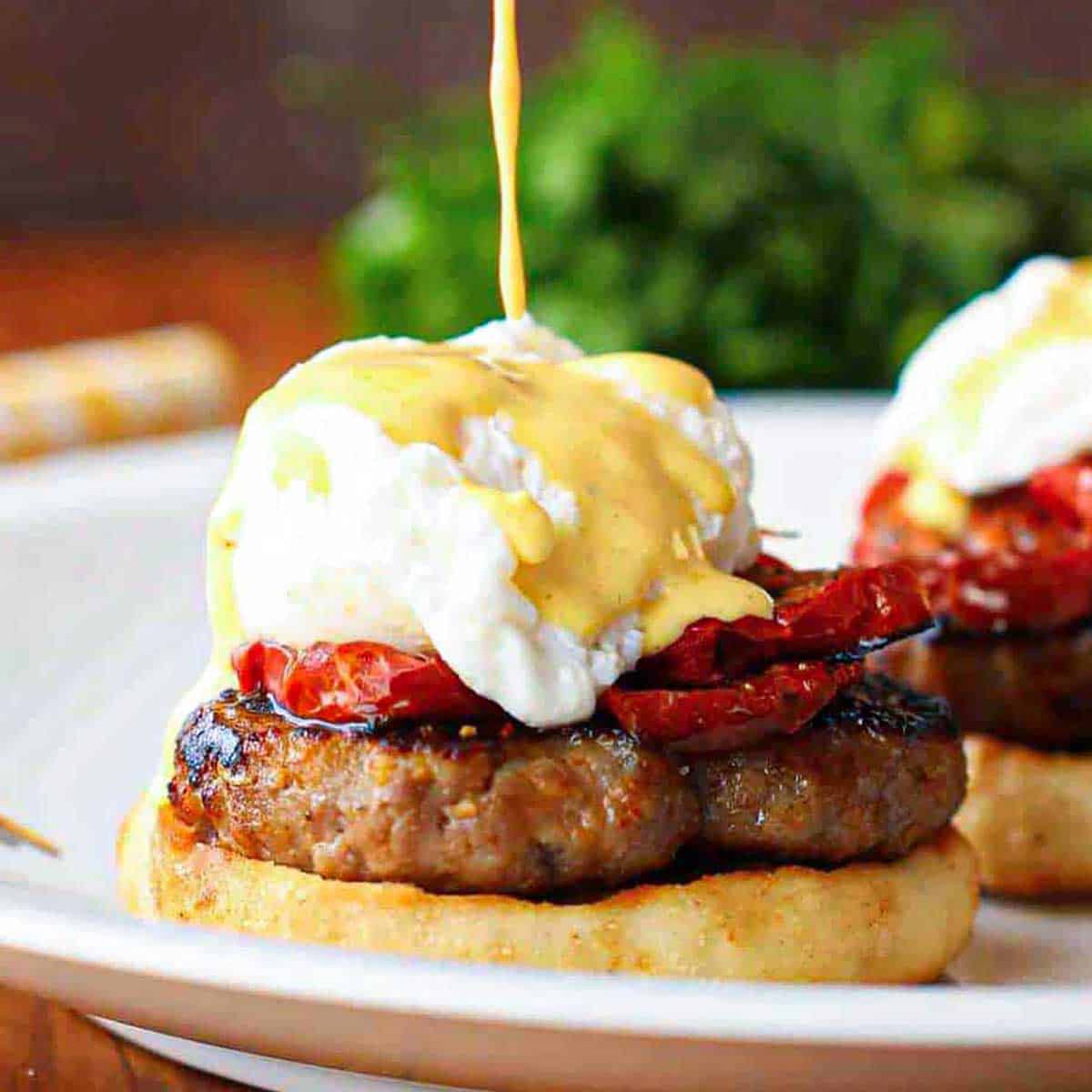 A thin drizzle of hollandaise sauce pouring onto a serving of Eggs Blackstone with Sausage on a white dinner plate.