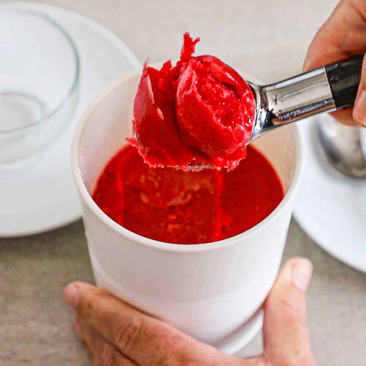 A person using an ice cream scooper to hold up a scoop of homemade raspberry sorbet from a pint ice cream holder filled with the same.