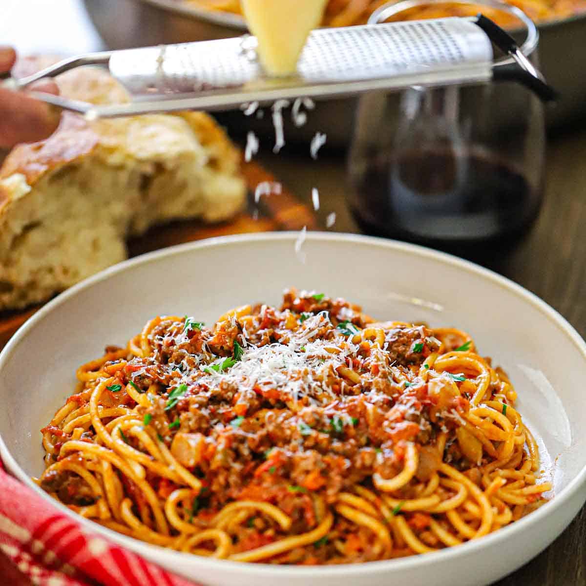 A person grating a block of Parmesan cheese on a microplane over a bowl of spaghetti bolognese in a shallow white pasta bowl.