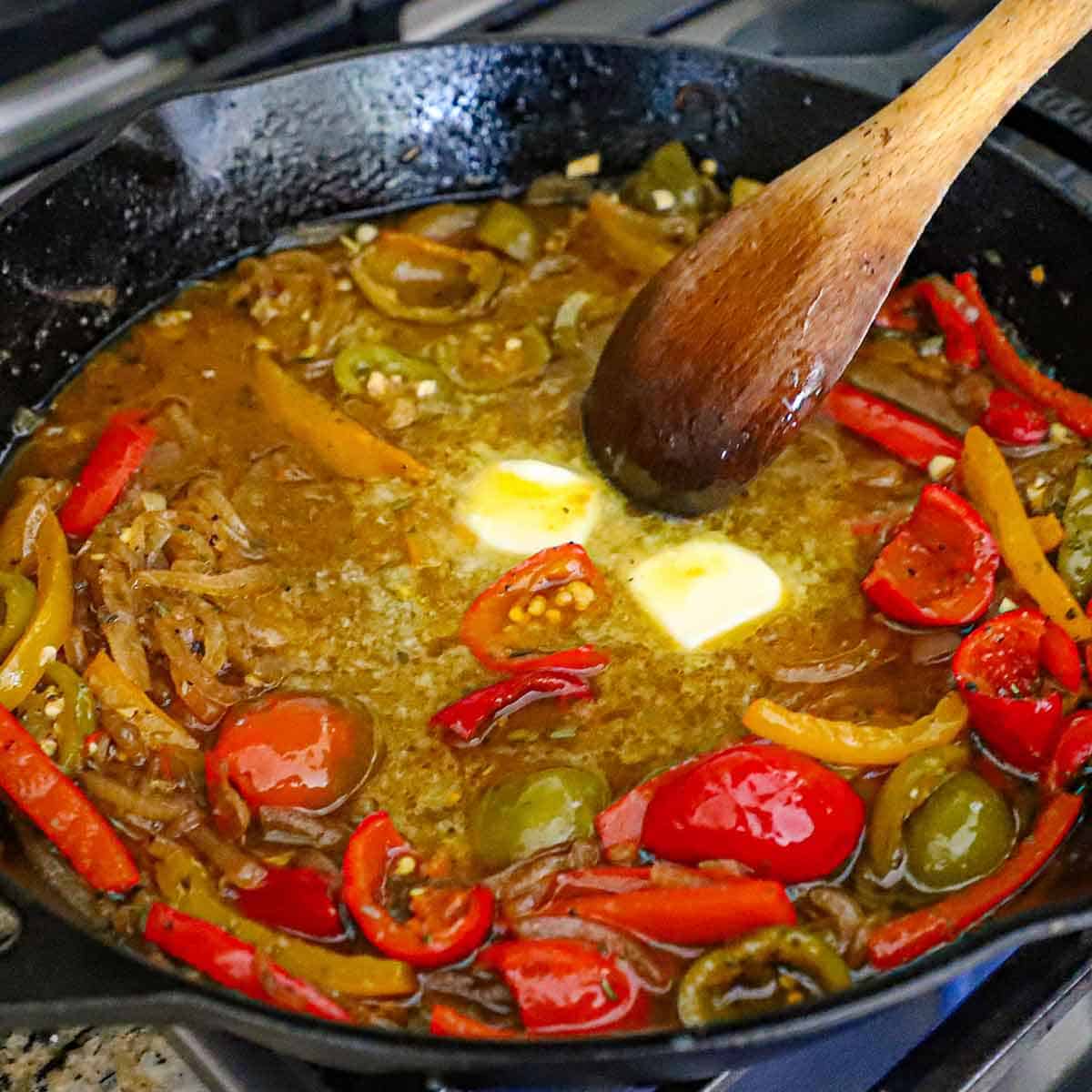 A person using a wooden spoon to stir two small pallets of butter that are melting in a simmering chicken scarpariello sauce in a skillet on a gas stove.