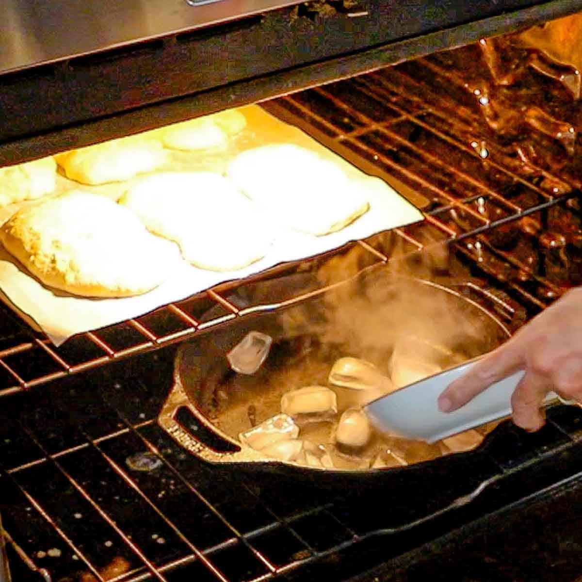 A person adding ice cubes to a large hot cast-iron skillet in a hot oven with a pizza steel on the rack above containing six ciabatta rolls that are about to be baked.