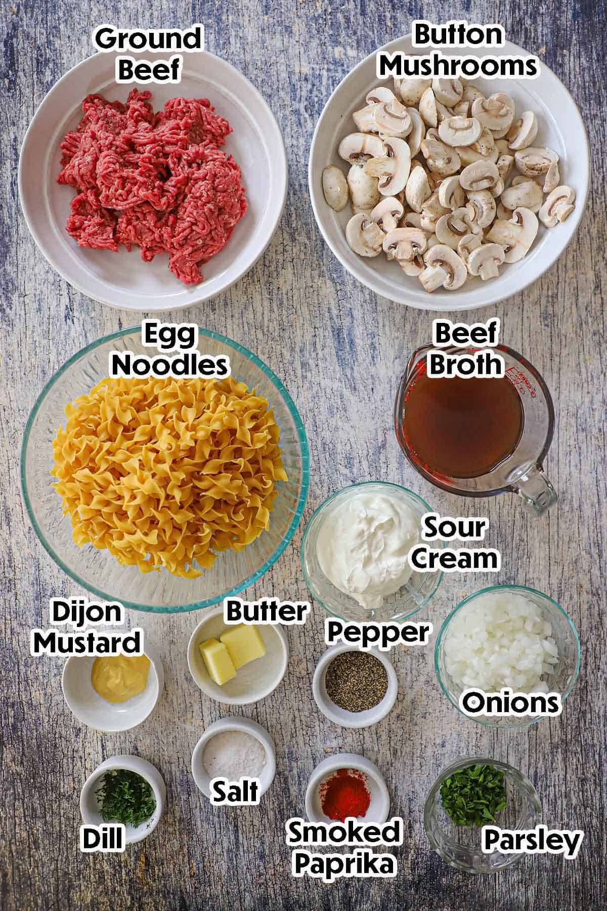 An arrangement of ingredients for Weeknight Beef Stroganoff on a grey-wooden background including ground beef, sliced mushrooms, beef broth, onions, egg noodles, butter, herbs, and seasonings.